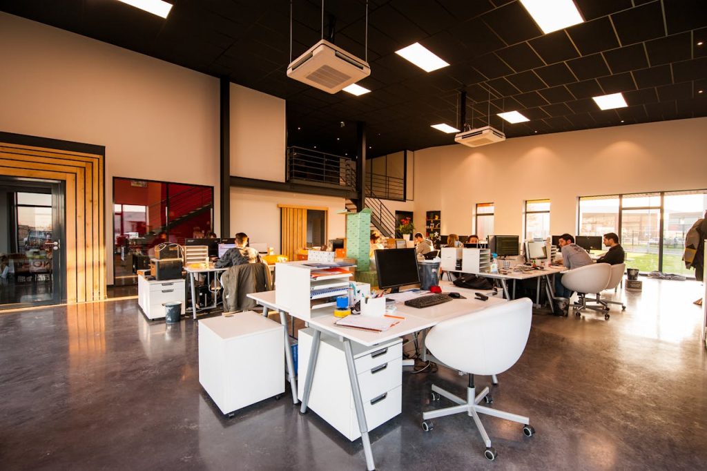 Modern open-plan office with white desks, employees working at computers, dark ceiling, and large windows with natural light.