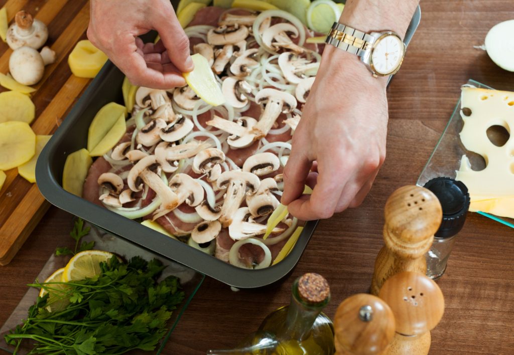 Hands layering sliced mushrooms and onions over raw meat in a baking pan with cheese, herbs, and oil nearby