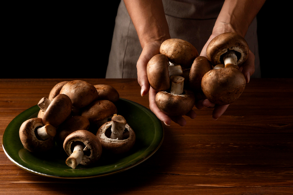 Hands holding fresh cremini mushrooms over a green plate piled with brown button mushrooms on a wooden table.